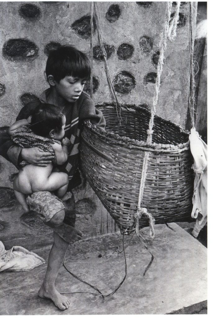 Black and white photo of a child holding a baby balanced on their leg while they sort out the baby's cradle.