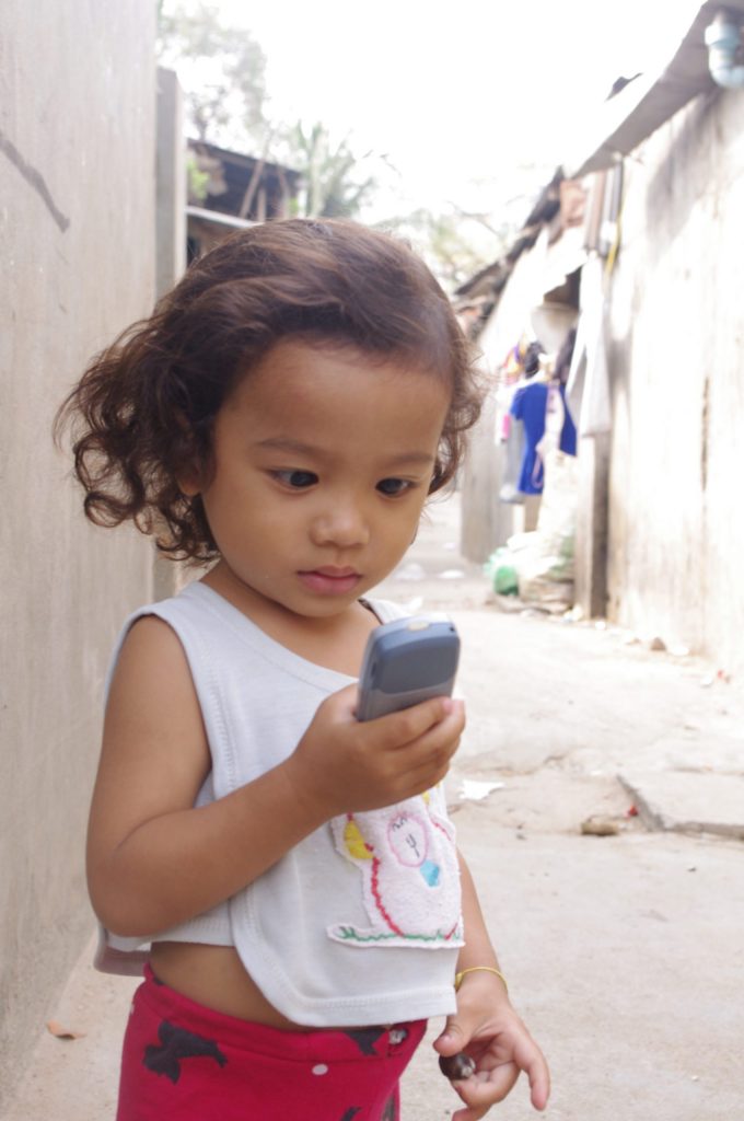 A toddler in a white sleeveless top stands in the street looking at a mobile phone.