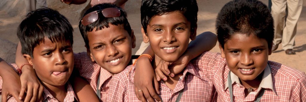 Four children stand together with their arms around each other. They are wearing red and white button-up shirts.