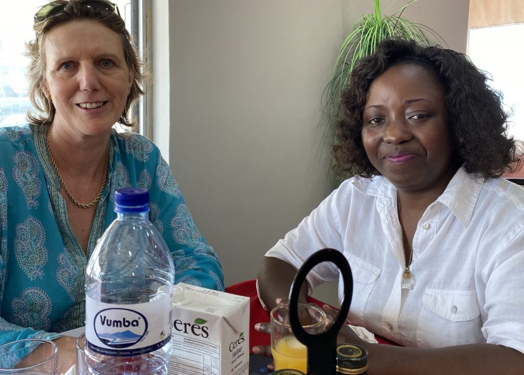 Clare (in a blue and white shirt) and Bibiche (in a white shirt) sit at a table with a red tablecloth with glasses of water and juice in front of them.