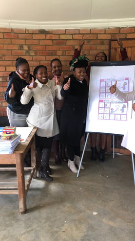 A group of teachers wave to the camera with two Children for Health posters visible on a flip chart next to them.